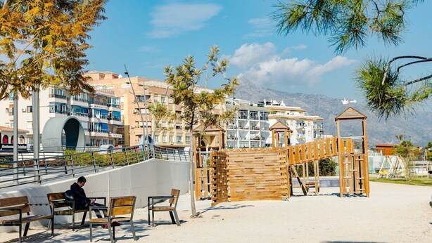 Un parque junto a la playa con bancos de madera, un área de juegos y edificios al fondo.