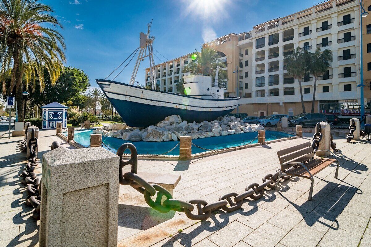 A large ship sculpture in a public square with a pool, surrounded by stone bollards and chains.