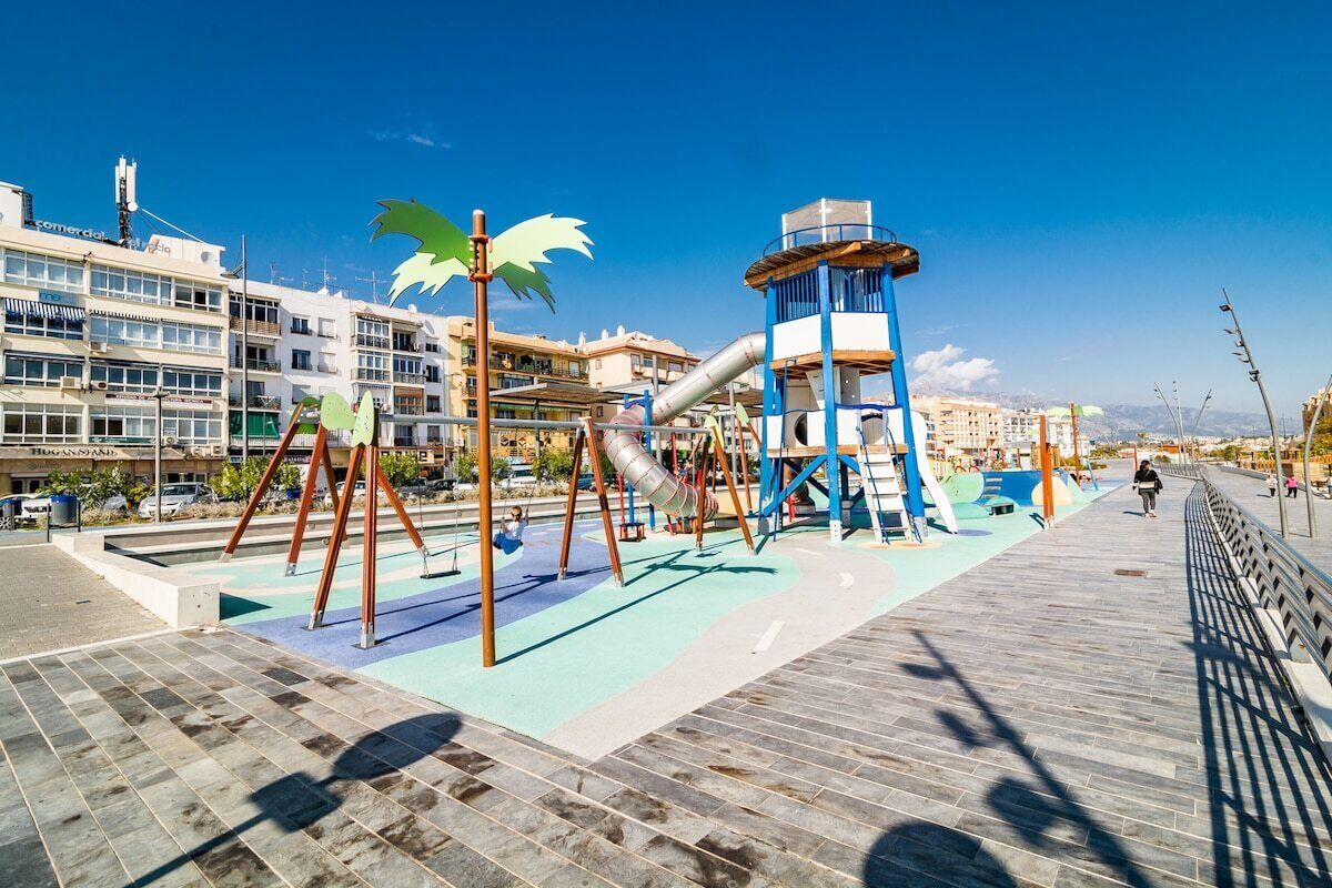 Ein Spielplatz mit Rutschen und Schaukeln, ein blaues und grünes Spielareal und eine Fußgängerbrücke mit Geländer.