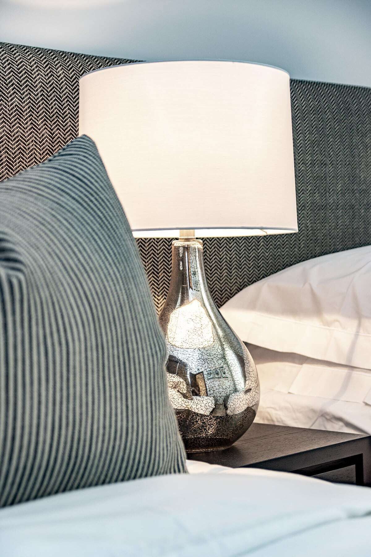 En säng med en vit kudde och en randig kudde, en metallampa med en vit lampskärm och en vägg med en tydlig struktur bakom.