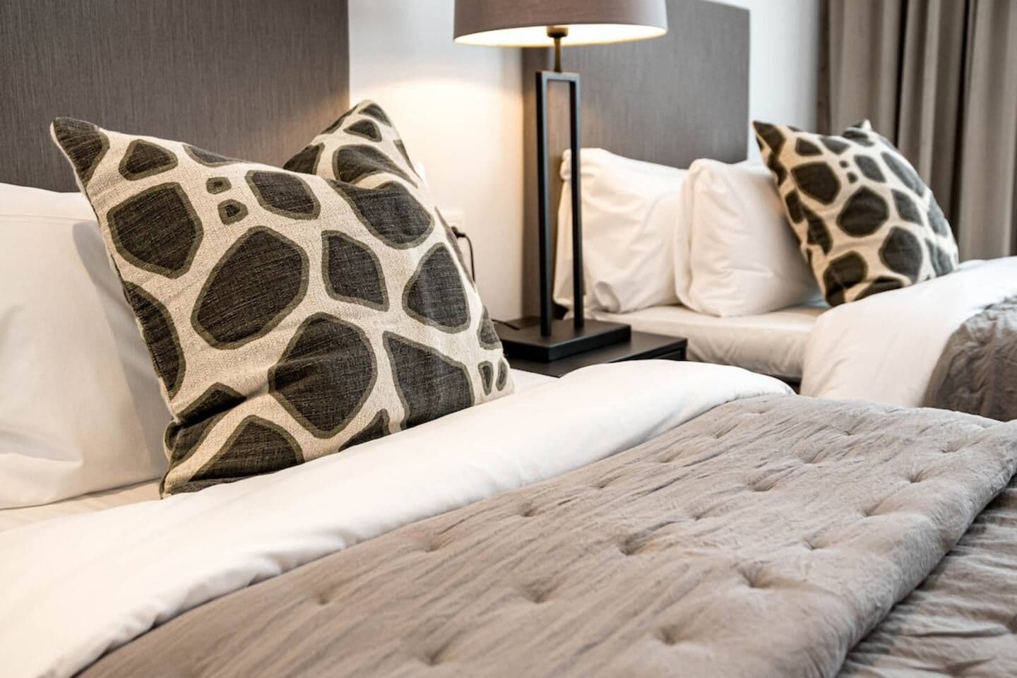 A hotel room with a bed, two pillows with a leaf pattern, a bedside lamp, and a grey blanket.