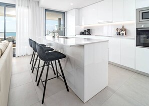 A modern kitchen with white cabinetry, a central island with bar stools, and a view of the sea through large windows. - Lme133a- Stunning Flat Close to Beach in Estepona (Estepona)