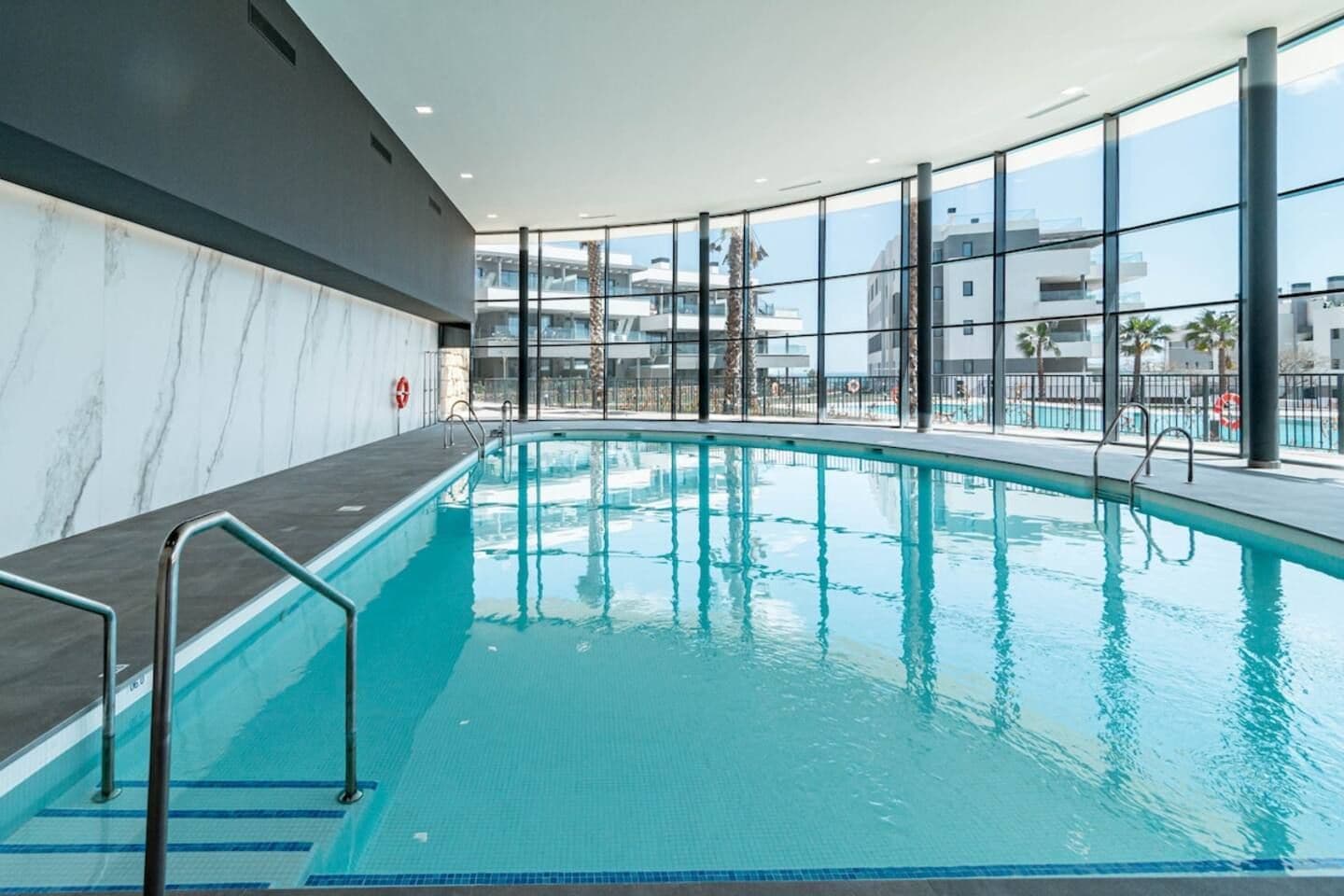 Une piscine intérieure avec des parois en verre transparent, un mur en marbre et une vue sur les bâtiments environnants.