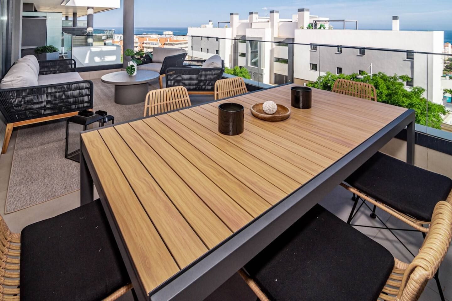 A modern outdoor dining area with a wooden table and chairs, a round coffee table, and a view of the cityscape.