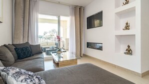 A modern living room with a sofa, a glass coffee table, and a TV mounted on the wall. There are decorative items on shelves and a view of the outside balcony. - Cpg- Perfect Holiday Home Close to Puerto Banus (Marbella)