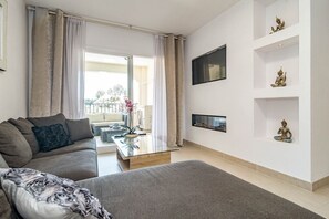 Een moderne woonkamer met een bank, een glazen salontafel en een aan de muur bevestigde televisie. Decoratieve objecten staan op planken en er is uitzicht op het balkon.