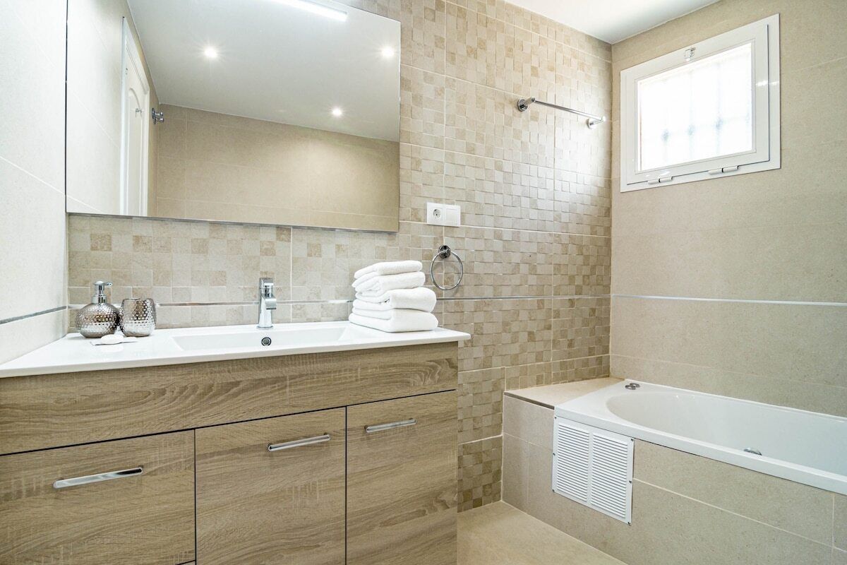 A modern bathroom with two sinks, a large mirror, a bathtub, and a window.