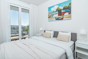 A bedroom with a large bed, two bedside tables, a painting on the wall, and a view of the city through a sliding door. - JG- Top Modern Apartment in Nueva Andalucia (Marbella)