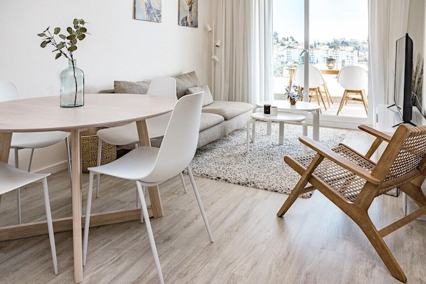 Un salon moderne avec une table de salle à manger ronde, des chaises blanches, un canapé et un fauteuil en bois. On peut voir un vase avec des feuilles vertes sur la table, et un téléviseur sur la droite.