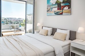 A modern hotel room with a large bed, a bedside table with a lamp, a view of the city through a large window, and a painting on the wall. - JG- Top Modern Apartment in Nueva Andalucia (Marbella)