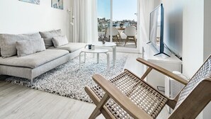 A modern living room with a sofa, coffee table, and a TV. There are two paintings on the wall, a balcony with a view, and a large window with curtains. - JG- Top Modern Apartment in Nueva Andalucia (Marbella)