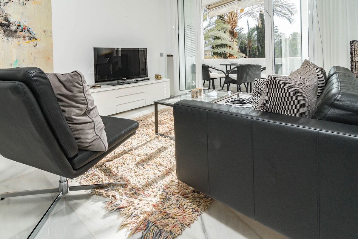 Una sala de estar moderna con un sofá de cuero negro, una mesa de centro de vidrio y un televisor sobre un mueble blanco.