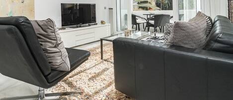 Una sala de estar moderna con un sofá de cuero negro, una mesa de centro de vidrio y un televisor sobre un mueble blanco.