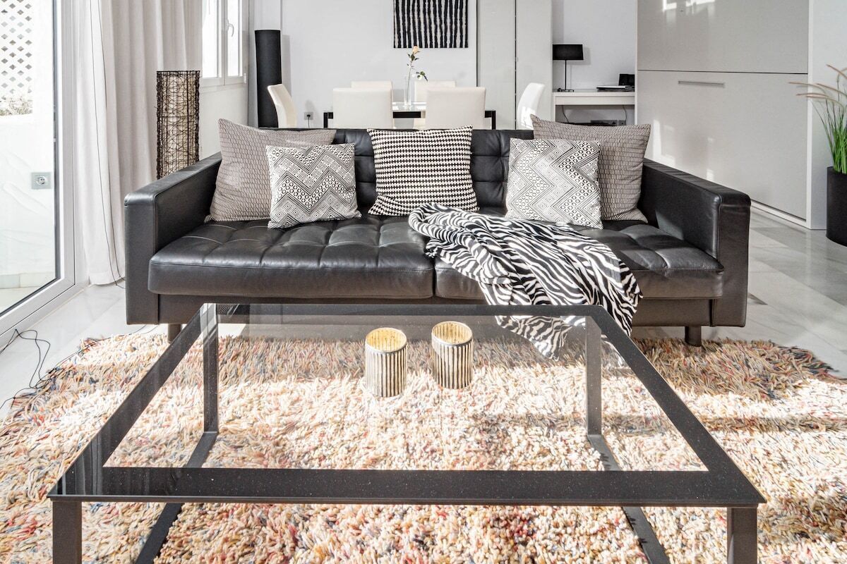 A modern living room with a leather sofa, patterned cushions, a glass coffee table, and a textured rug.