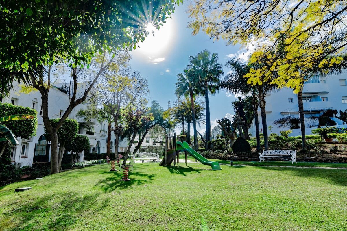 Un parque con un tobogán verde, bancos y palmeras.