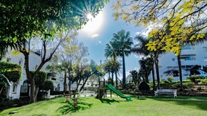Un parque con un tobogán verde, bancos y palmeras.