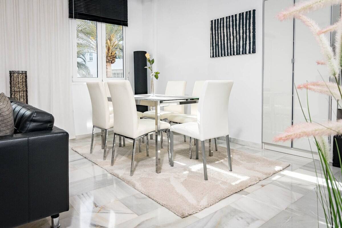 A modern dining area with a glass table and white chairs, a black sofa, and a decorative vase with flowers.