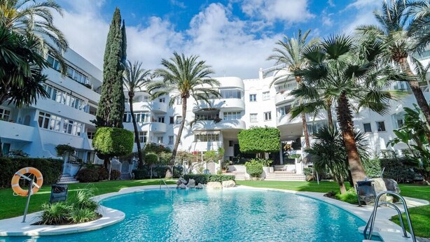 Piscina rodeada de palmeras y un edificio blanco con balcones.
