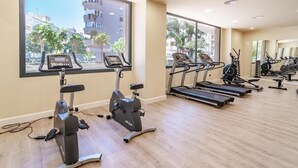 Ein Fitnessraum mit Cardiogeräten, darunter Laufbänder und Ergometer, großen Fenstern mit Blick auf Palmen und Gebäude sowie Holzböden.