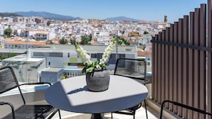 Ein Balkon mit einem runden Tisch und zwei Stühlen, mit Blick auf eine Stadtlandschaft mit Gebäuden und Bergen.