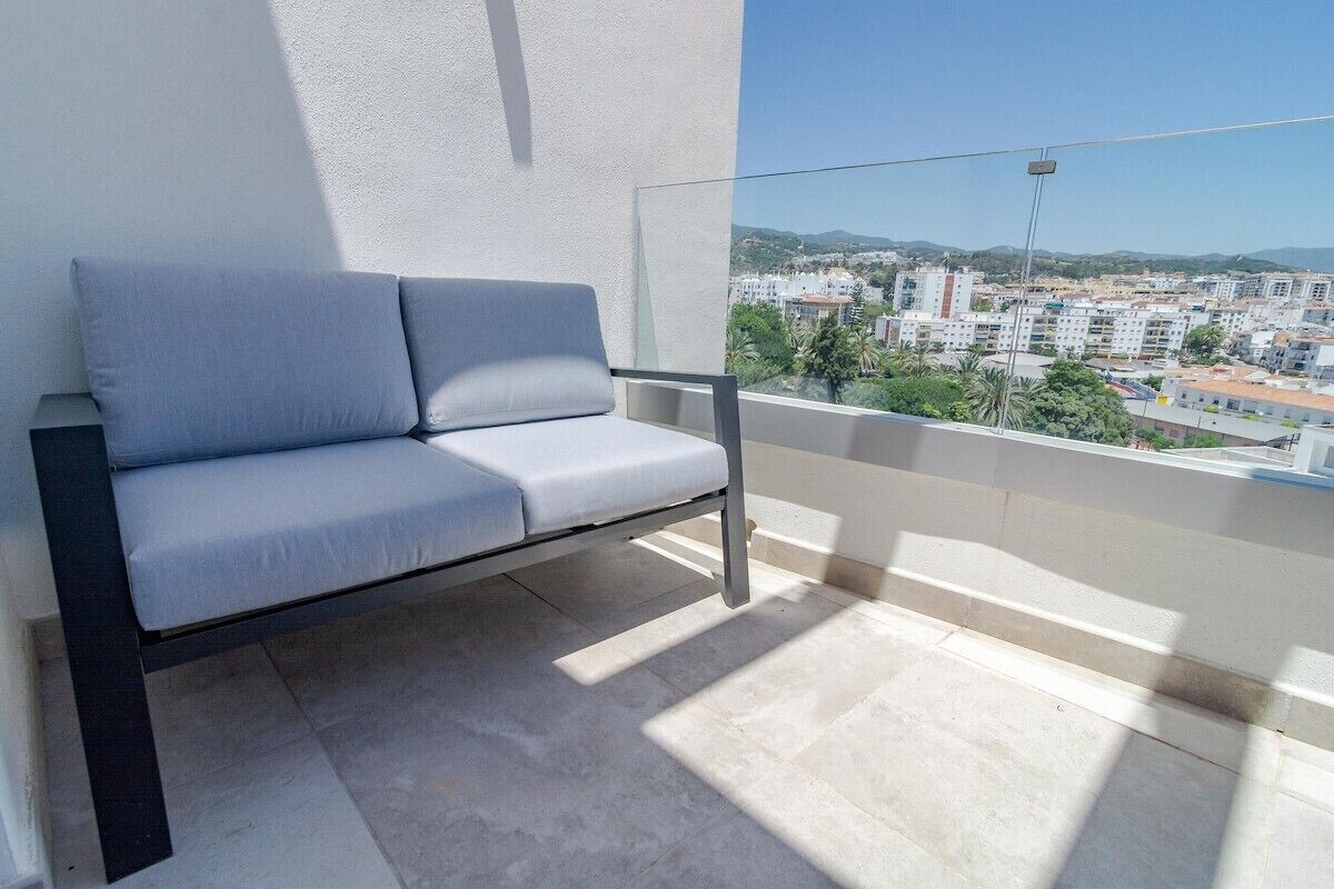A balcony with a grey sofa, a clear view of a cityscape, and a white wall.
