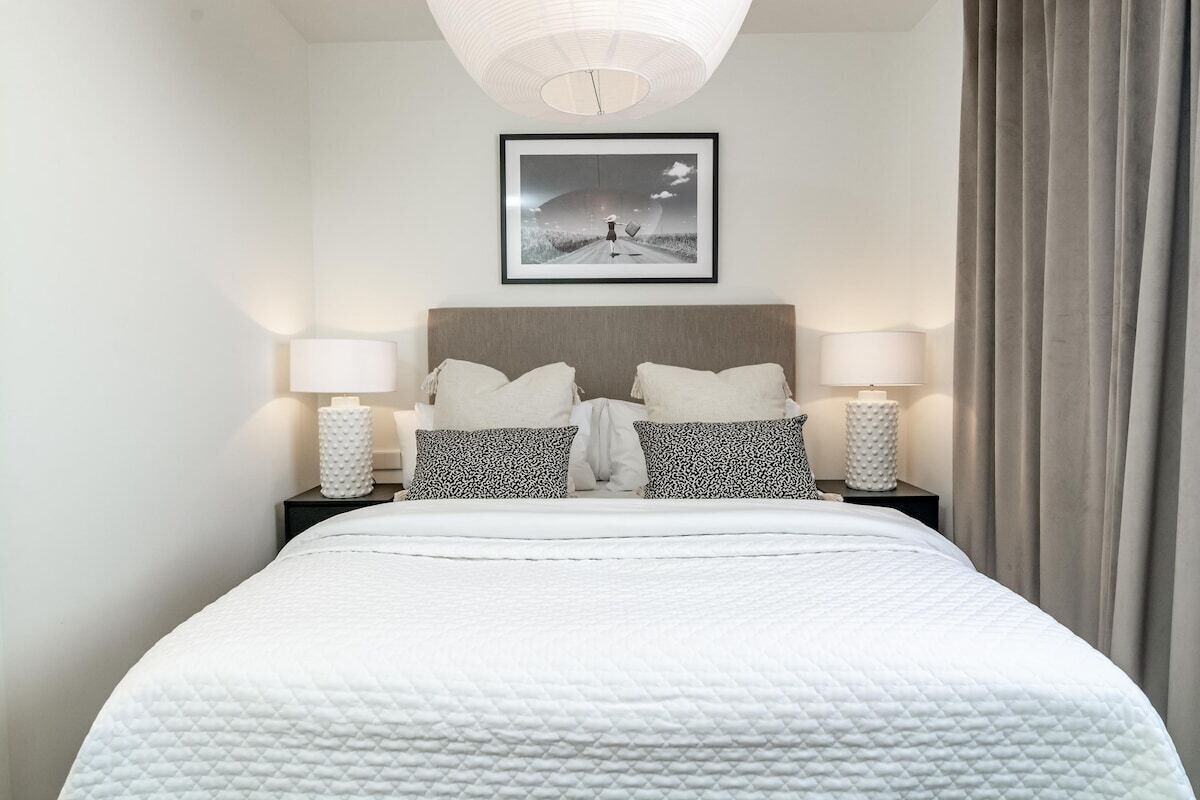Un letto rifatto con coperta bianca trapuntata, due comodini con lampade, un quadro incorniciato al muro e una finestra con tende.
