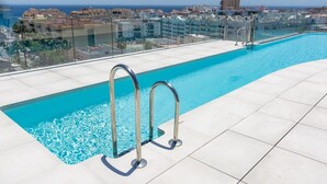 Uma piscina infinita no terraço, com água azul cristalina, escadas de metal e vista para a cidade e o mar ao fundo.