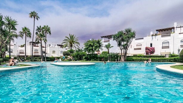 A large outdoor swimming pool surrounded by palm trees and a white building with arched windows. - LVB - Casa Linda Vista by Roomservices (Marbella)