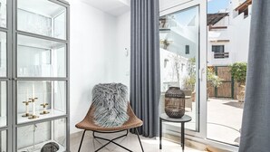 A modern interior with a glass display cabinet, a cushioned chair, a small table with a decorative vase, and a view of a residential area through a large window. - LVB - Casa Linda Vista by Roomservices (Marbella)