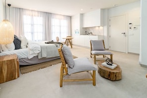 Ein modernes Hotelzimmer mit einem Bett, zwei Stühlen, einem runden Holztisch und einer Küchenzeile im Hintergrund.