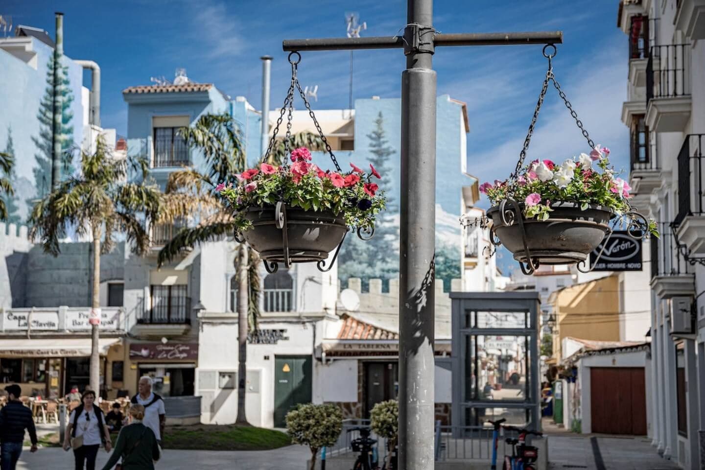 Une scène de rue avec des jardinières suspendues, une zone piétonne et des bâtiments portant des enseignes.