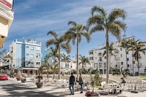 A sunny outdoor cafe area with palm trees, outdoor seating, and buildings in the background. - A2- Seaclub Suites by Roomservices (Estepona)