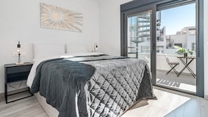 A modern bedroom with a large bed, a bedside table with a lamp, a wall-mounted abstract art piece, and a sliding glass door leading to a balcony with a table and chairs. - Eg54a - Luxury Penthouse With Private sun Terrace (Estepona)
