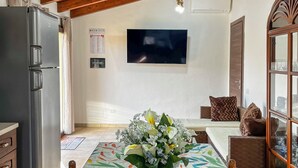 Een eethoek met een lange tafel, stoelen, een koelkast, een televisie en een decoratieve plant.