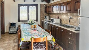 Une cuisine avec des meubles de rangement en bois, une table à manger recouverte d’une nappe à motifs floraux et une fenêtre avec des rideaux.