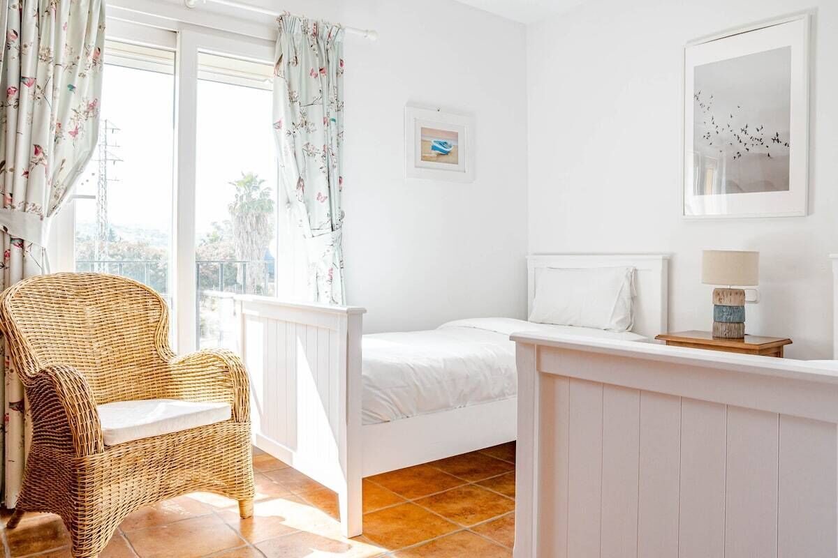 Une chambre à coucher avec un lit, une chaise en osier, une fenêtre avec des rideaux et un tableau encadré accroché au mur.