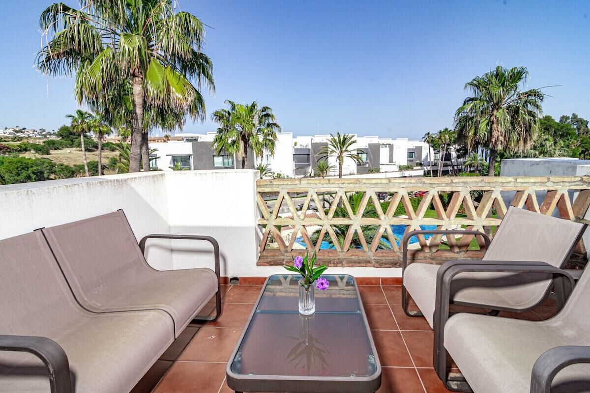 Un balcon avec deux canapés de couleur beige, une table avec un plateau en verre et un vase rempli de fleurs. On aperçoit des palmiers et une piscine à l’arrière-plan.