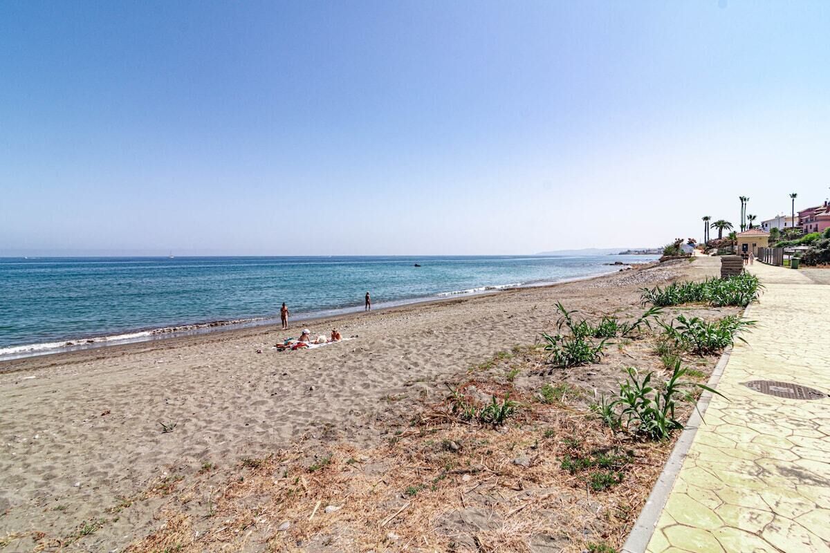 Una playa con un paseo marítimo empedrado, algunas personas y edificios al fondo.