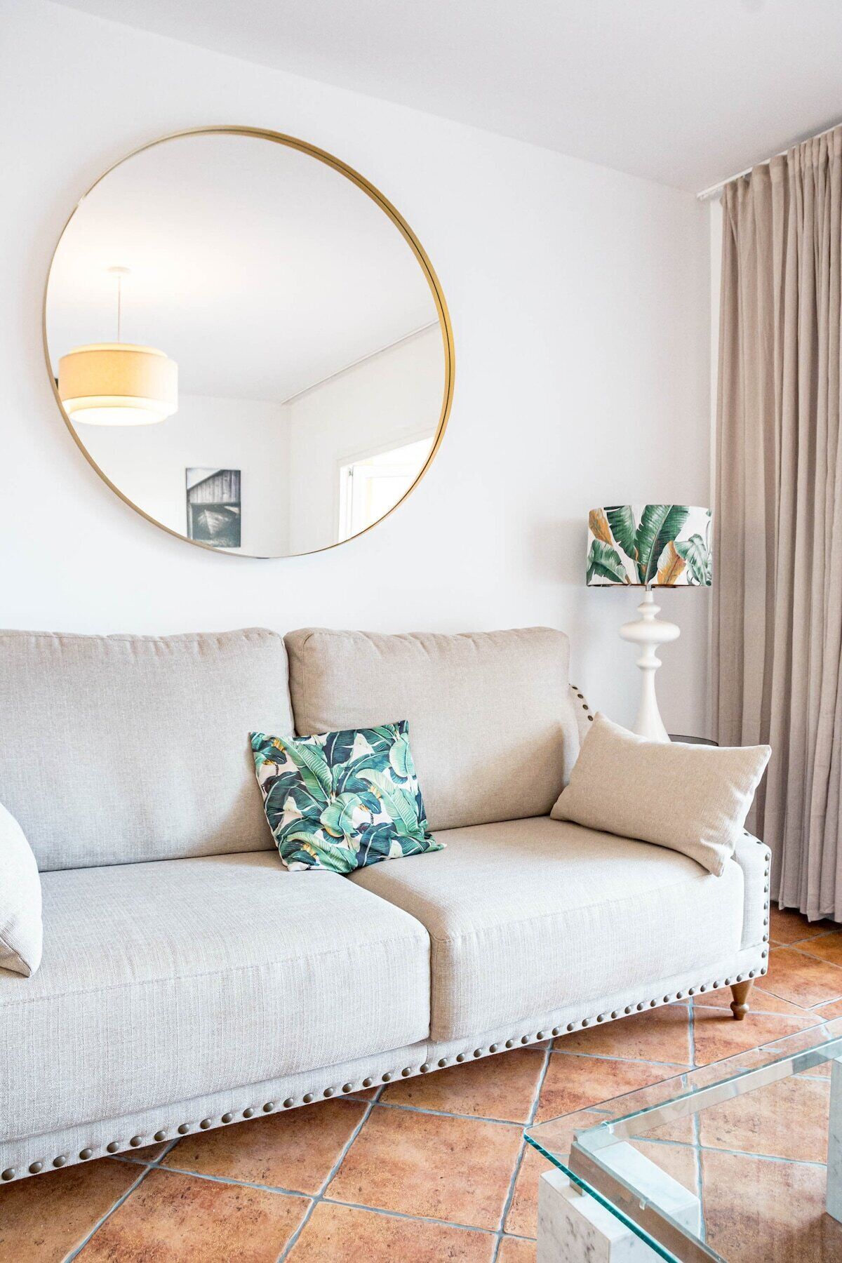 En moderne stue med en lysegrå sofa, to dekorative puder, et rundt spejl, en hvid lampe med en plante, og et glasbord.