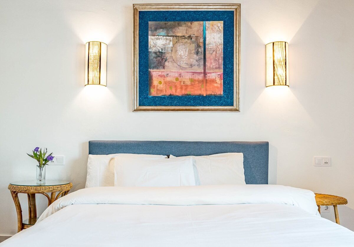 A hotel room with a bed, a framed abstract painting, two wall-mounted lights, and a small table with a vase of flowers.