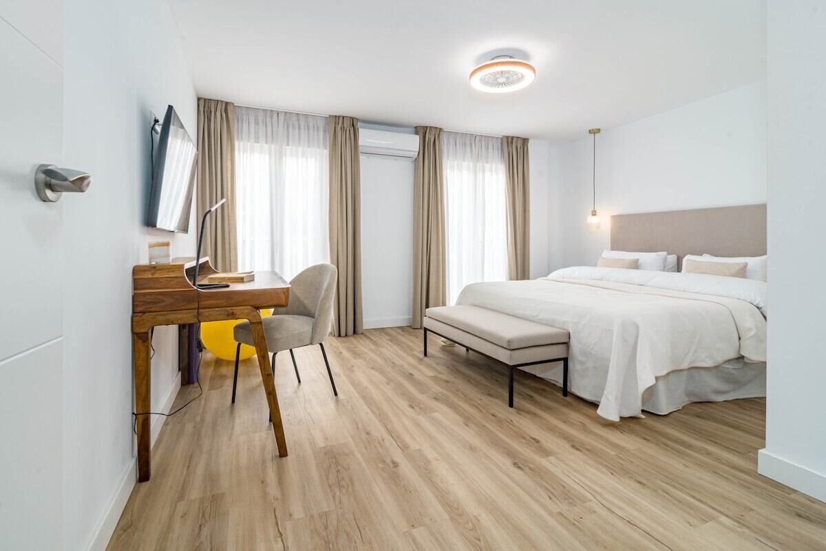 Une chambre d’hôtel moderne avec un bureau en bois, un fauteuil confortable, un lit avec du linge de maison blanc et une grande fenêtre avec des rideaux.
