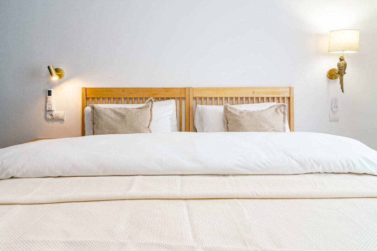A neatly made bed with a wooden headboard, two bedside lamps, and a wall-mounted thermostat.