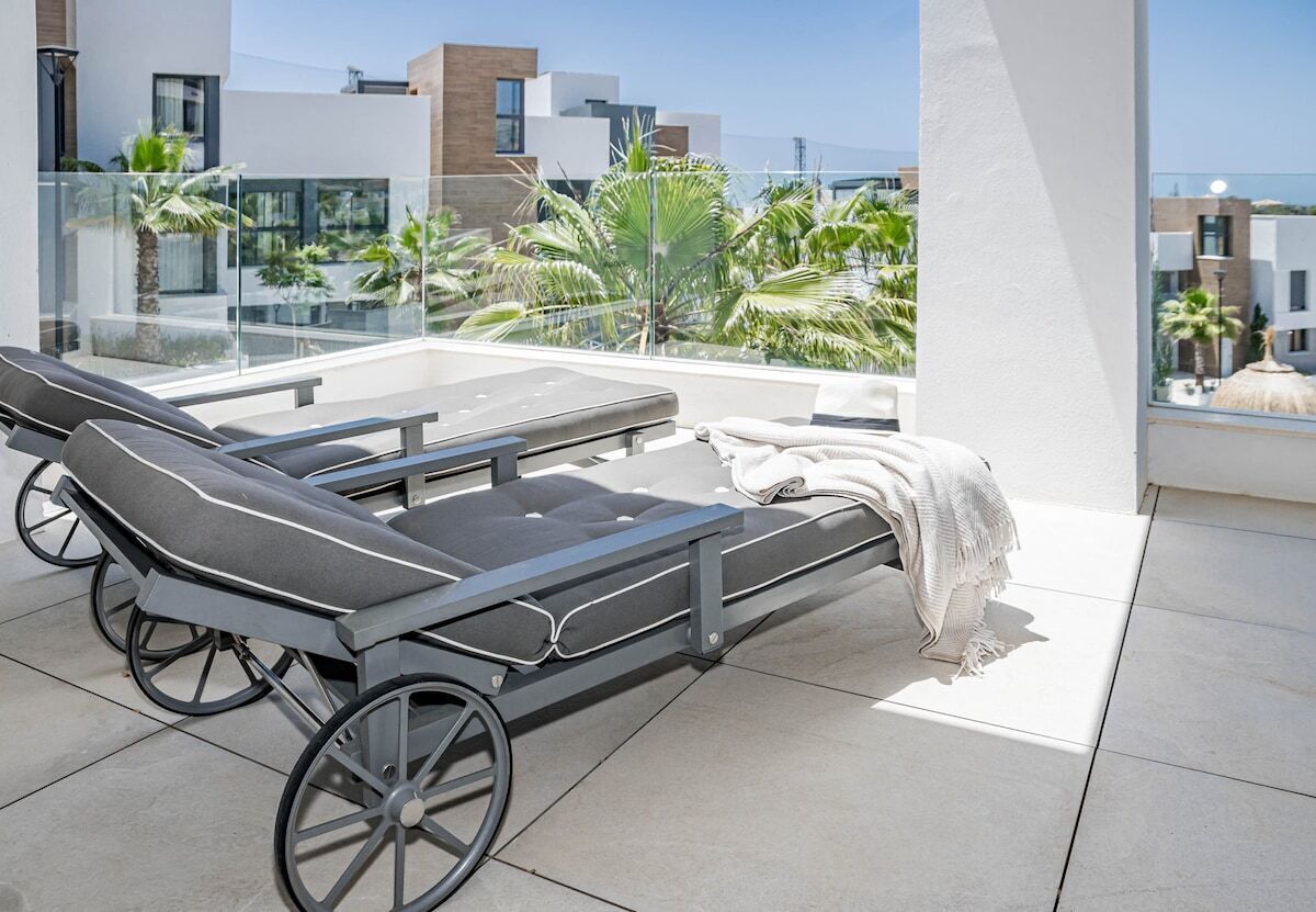 A modern outdoor lounge area with a reclining chair and a small table, overlooking a cityscape with palm trees and modern buildings.