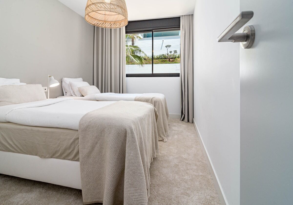 Quarto moderno com uma cama grande, duas mesas de cabeceira, uma luminária e uma janela com cortinas, oferecendo vista para o mar.