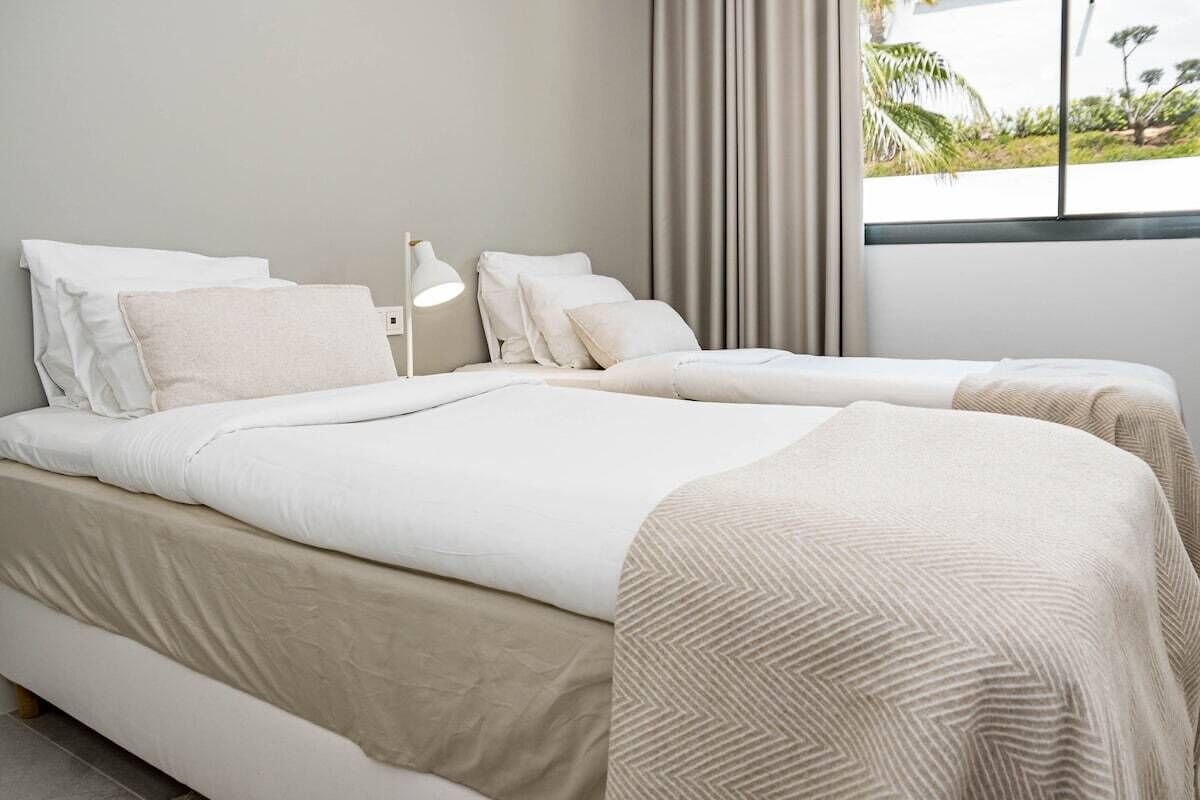 A modern hotel room with two beds, a large window with curtains, and a view of greenery outside.