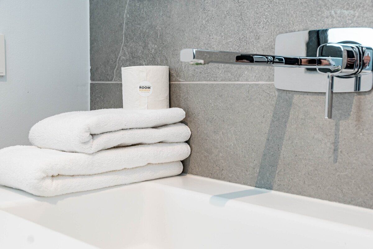 Une salle de bain avec un lavabo blanc, une pile de serviettes blanches et un distributeur de papier toilette portant l’inscription « SERVICES DE L’HÔTEL ».