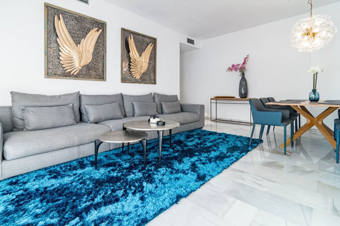 Een moderne woonkamer met een grijze bank, twee ronde salontafels, een blauw pluchen vloerkleed en een eetgedeelte met een houten tafel en stoelen.