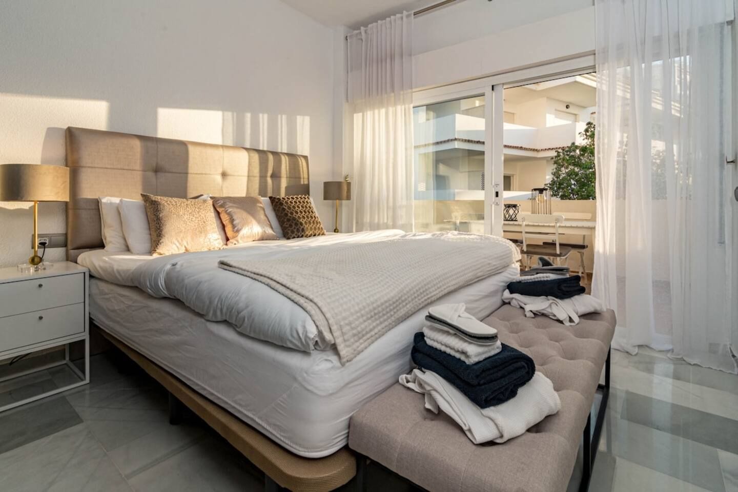 Une chambre moderne avec un grand lit, des lampes de chevet, un banc avec des serviettes et une vue sur l’extérieur grâce à de grandes fenêtres.
