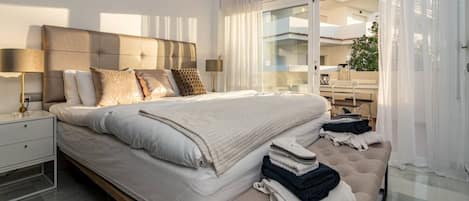 A modern bedroom with a large bed, bedside lamps, a bench with towels, and a view of the outdoors through large windows.