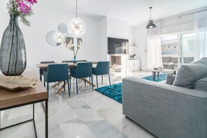 Uma sala de estar moderna com um sofá cinzento, uma área de jantar com mesa e cadeiras de madeira, e uma cozinha com uma televisão.